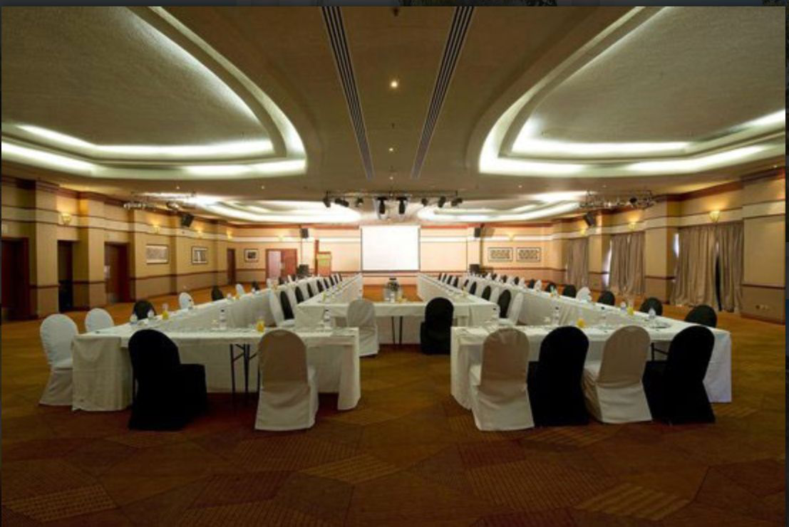 Conference hall arranged in a horseshoe of white-clothed tables facing a projection screen